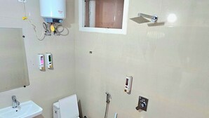 A small bathroom with a sink, toilet, and shower. There is a white heater mounted on the wall, a mirror, and a small shelf with toiletries. - Hotel Trident Dwarka (New Delhi)