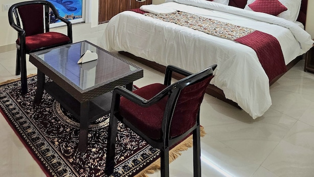 A hotel room with a bed, a desk, two chairs, a ceiling fan, an air conditioning unit, and a large poster of a building on the wall. - Hotel Trident Dwarka (New Delhi)