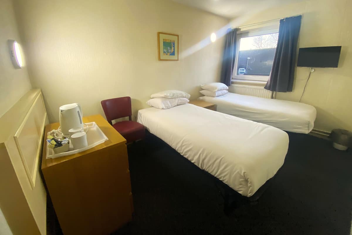 A hotel room with two single beds, a wooden desk with a coffee maker, a red chair, and a window with curtains.