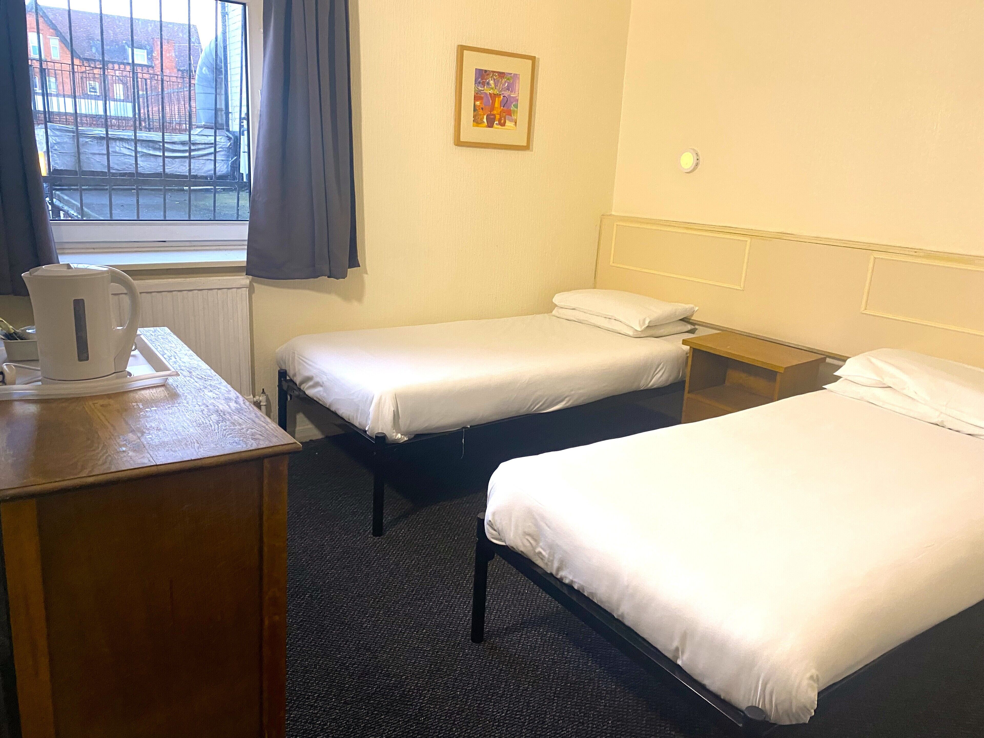 A hotel room with two single beds, a small desk with a kettle, a framed picture on the wall, and a window with curtains.