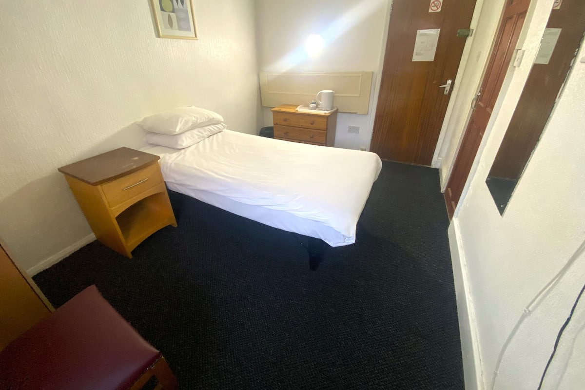 A small, neatly arranged hotel room with a single bed, a wooden nightstand, a small table with a cup, and a red chair.