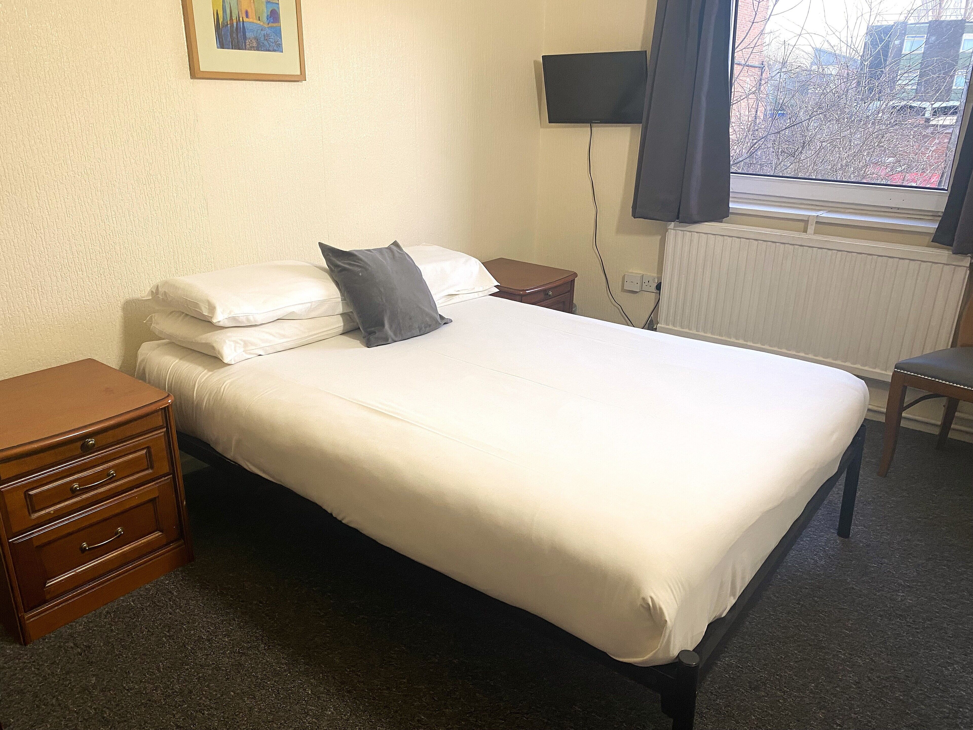 A neatly made bed with white linens and a gray pillow, a wooden nightstand with drawers, a small wooden dresser, a chair, a flat-screen TV mounted on the wall, and a window with a view of buildings outside.