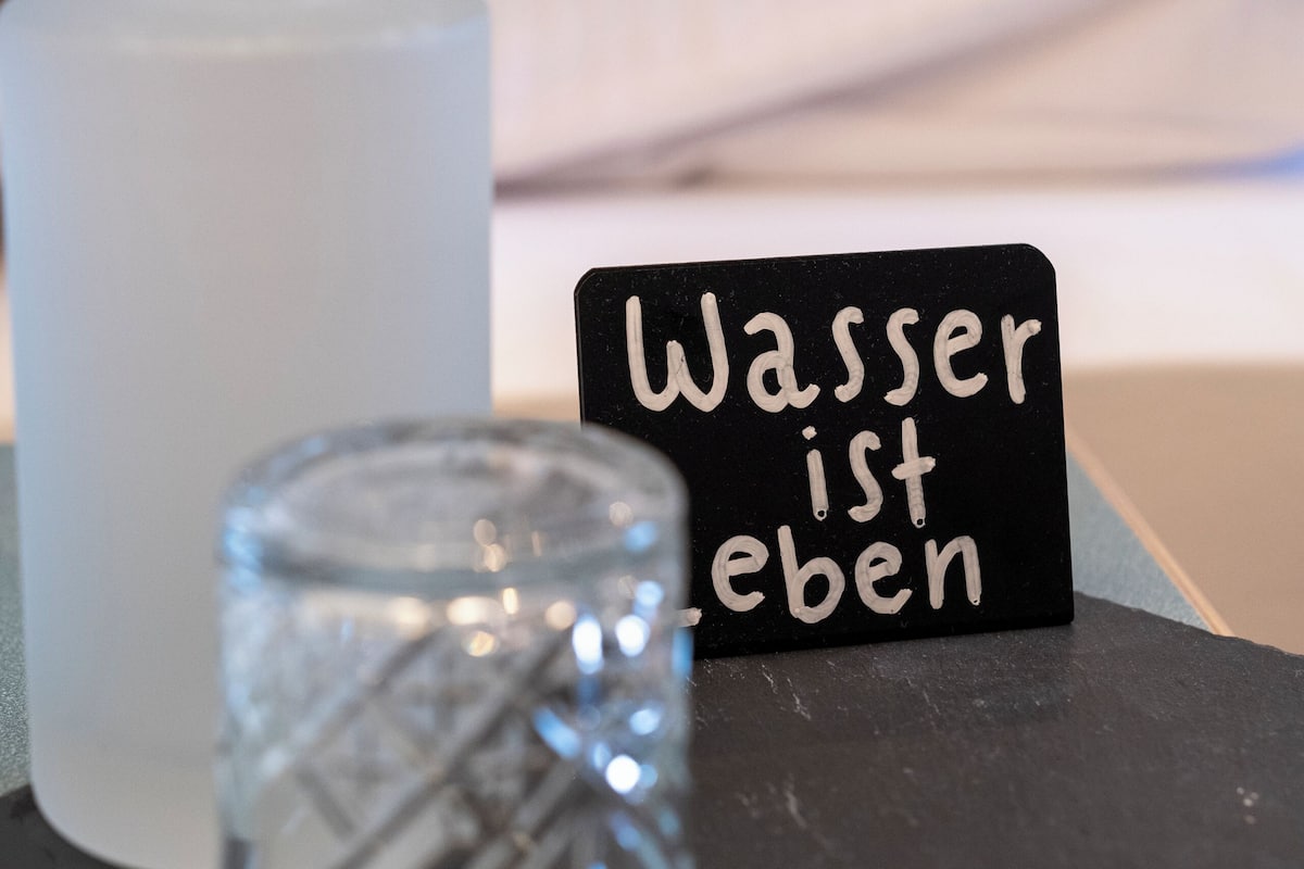 Ein Bett mit einem gelben Kopfkissen, ein schwarzes Schild mit weißer Schrift, eine weiße Flasche und ein klares Glas.