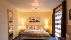 Habitación de hotel con una cama grande, dos mesitas de noche con lámparas, un espejo, un cuadro en la pared y una ventana con cortinas.