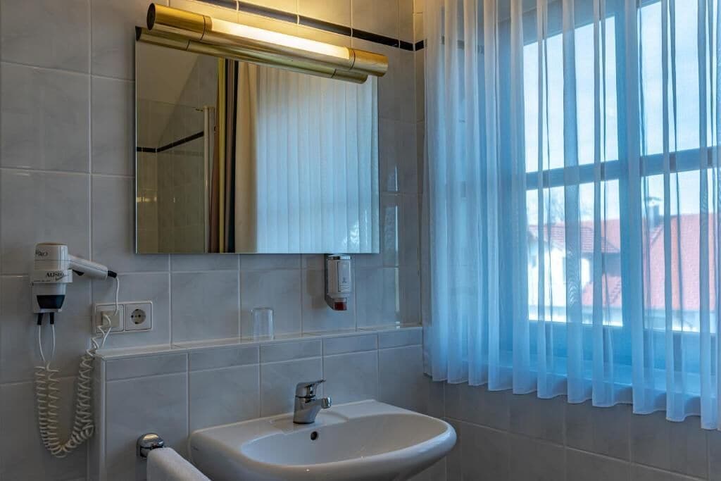 A bathroom with a white sink, a mirror, a hairdryer, and a window with sheer curtains.