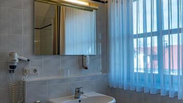 A bathroom with a white sink, a mirror, a hairdryer, and a window with sheer curtains.