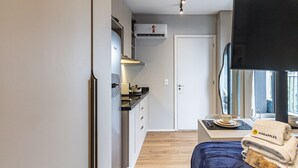 Et lille værelse med tekøkken, seng med blåt sengetæppe, fjernsyn og et vindue med gardiner.