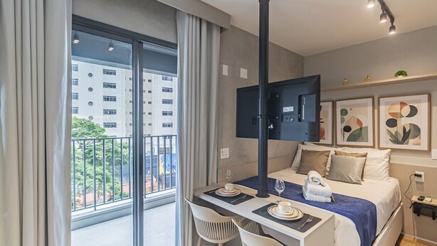 Et moderne hotelværelse med balkon, seng med blåt sengetæppe, skrivebord med stol og kunst på væggen.