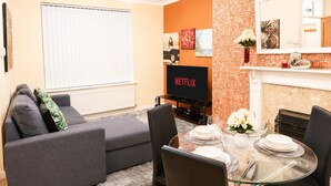 A living room with a grey sofa, a dining table set for four, a TV, and a brick wall. - Stunning 3-Bed House in the Heart of Newport, Sleeps 7 (Newport)
