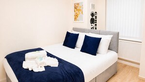 A neatly made bed with a dark blue bedspread, white linens, and two dark blue pillows. There is a folded towel on the bed. The room has light-colored walls, a window with blinds, and a radiator. - Stunning 3-Bed House in the Heart of Newport, Sleeps 7 (Newport)