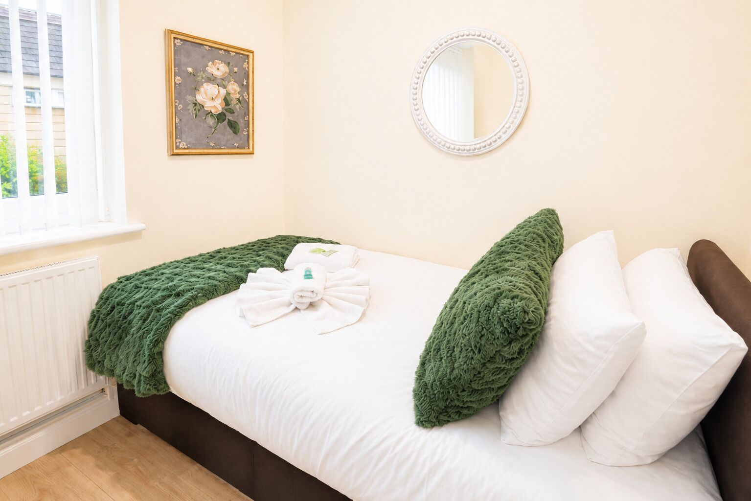 A bedroom with a bed, green throw, white pillows, a round mirror, and a floral painting on the wall.