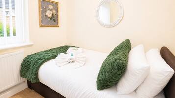 A bedroom with a bed, green throw, white pillows, a round mirror, and a floral painting on the wall.