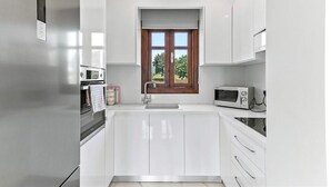Een moderne keuken met witte kasten, een inbouwoven, een magnetron en een raam met uitzicht op groen.