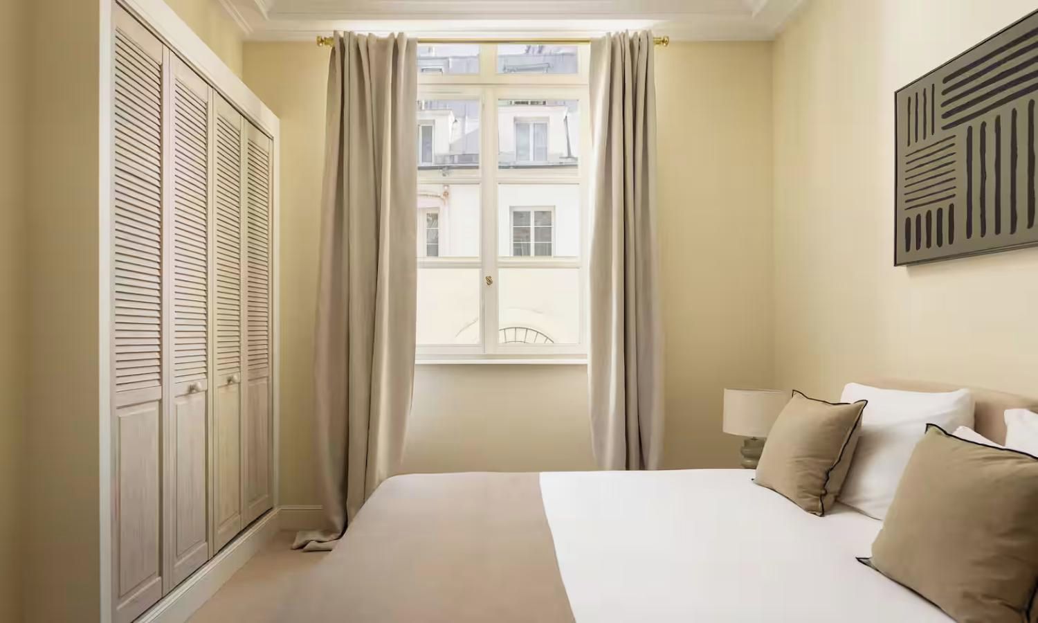 A hotel room with a bed, a window with curtains, a wall-mounted light, and a wardrobe.