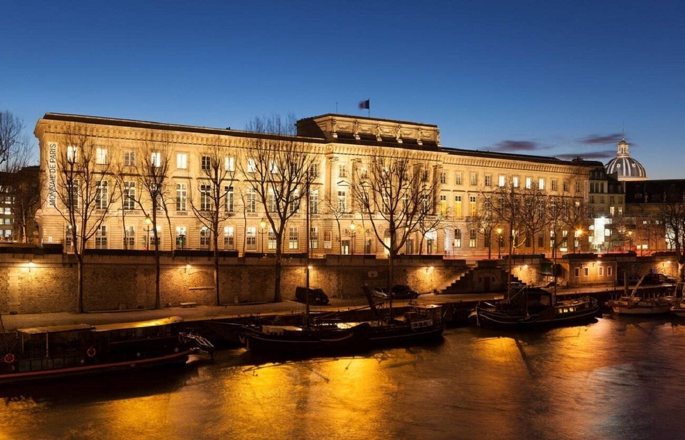 Un bâtiment historique à la façade classique, doté d’un dôme imposant et situé en bord de rivière, où des bateaux sont amarrés.