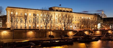Un bâtiment historique à la façade classique, doté d’un dôme imposant et situé en bord de rivière, où des bateaux sont amarrés.