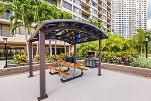 A covered picnic table with a grill in an outdoor area with palm trees and a high-rise building in the background. - Honolulu Condo Stay w/ Balcony: Walk to Beaches! (Honolulu)