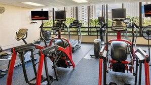 Ein Fitnessraum mit Cardiogeräten, einem Fernseher und Blick auf hohe Gebäude.