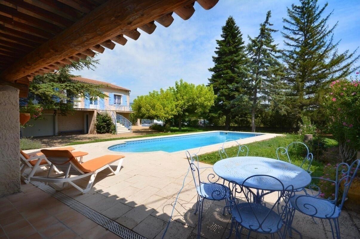 Zona de piscina con sillones de descanso y una mesa, rodeada de follaje y árboles.