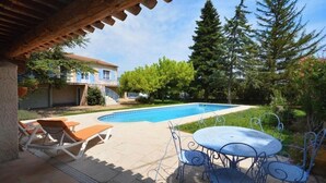 Una piscina amb gandules i una taula, envoltada de vegetació i un edifici al fons.