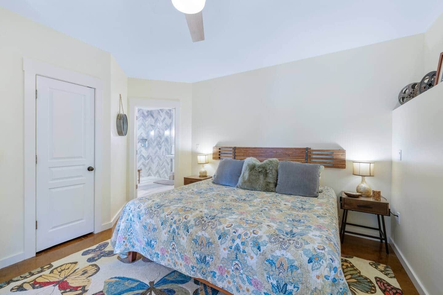 Un dormitorio con cama, una puerta, una mesita de noche, una lámpara y una alfombra estampada.