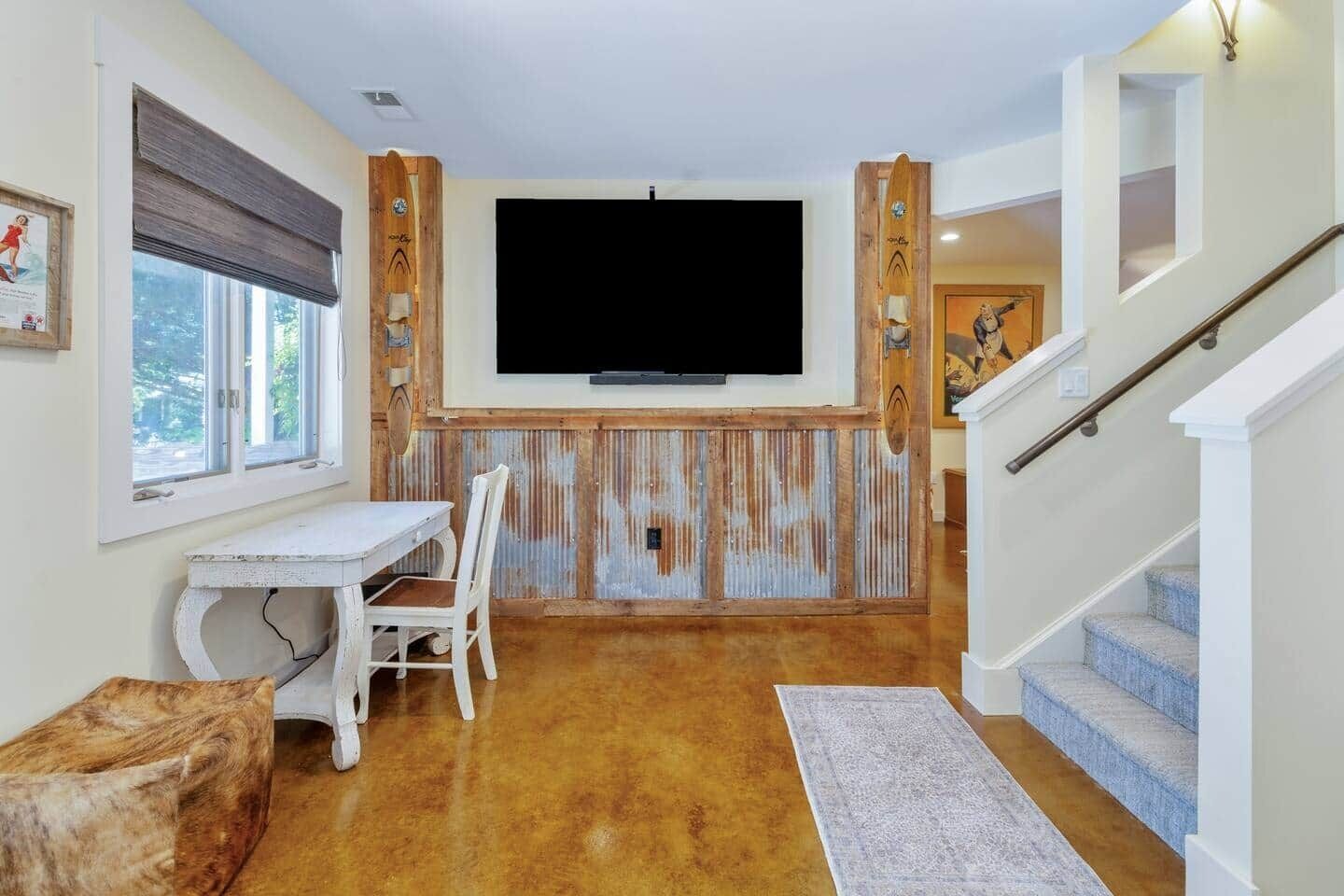 A living room with a TV mounted on a wooden wall, a dining table with chairs, a staircase, and a window.