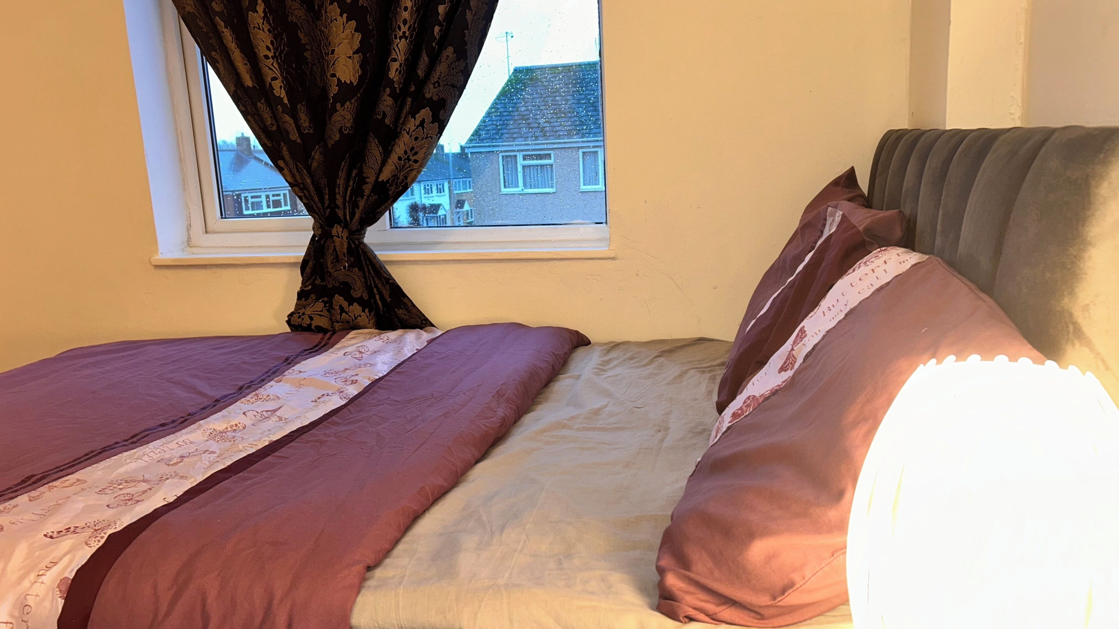 Un dormitorio con cama, una ventana con cortinas y vista a una casa.