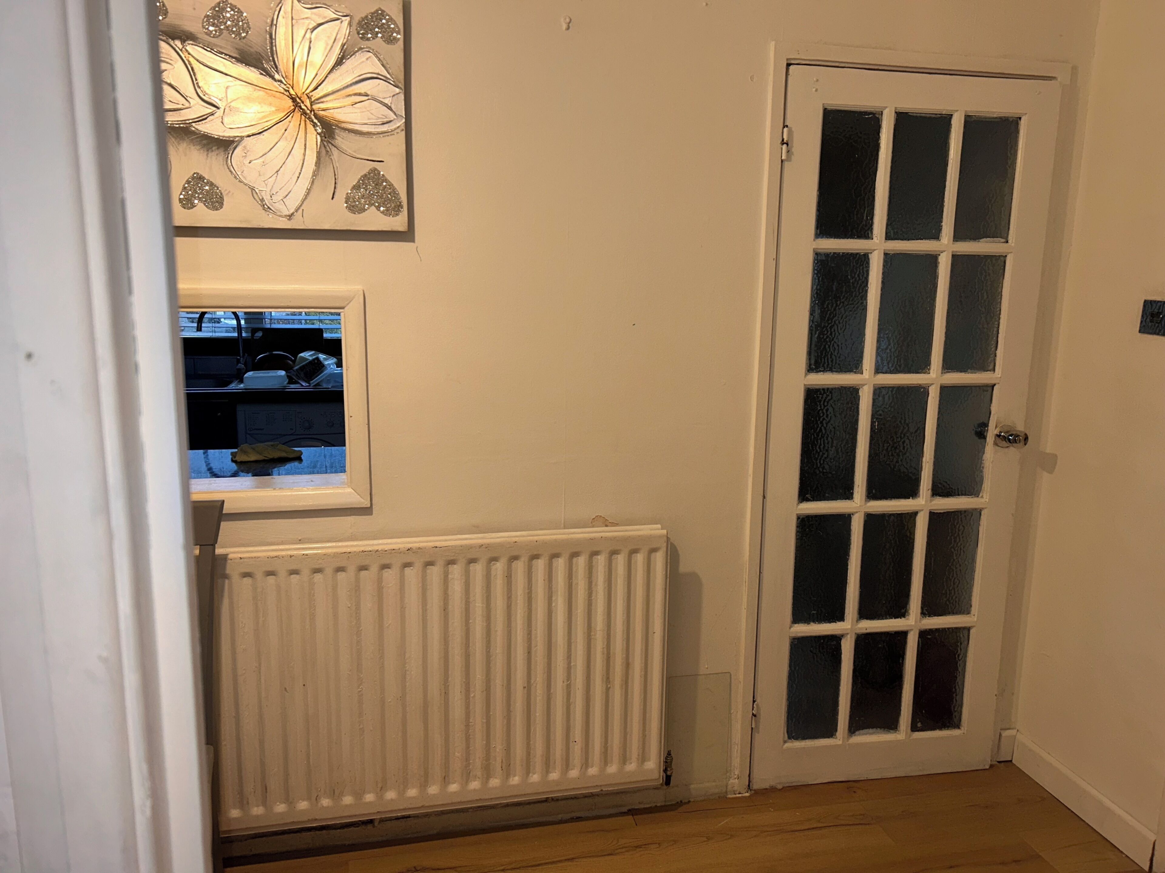 Ein Zimmer mit weißem Heizkörper, einer weißen Tür mit Glasfeldern und einem Fenster mit Blick auf eine Küchenzeile.