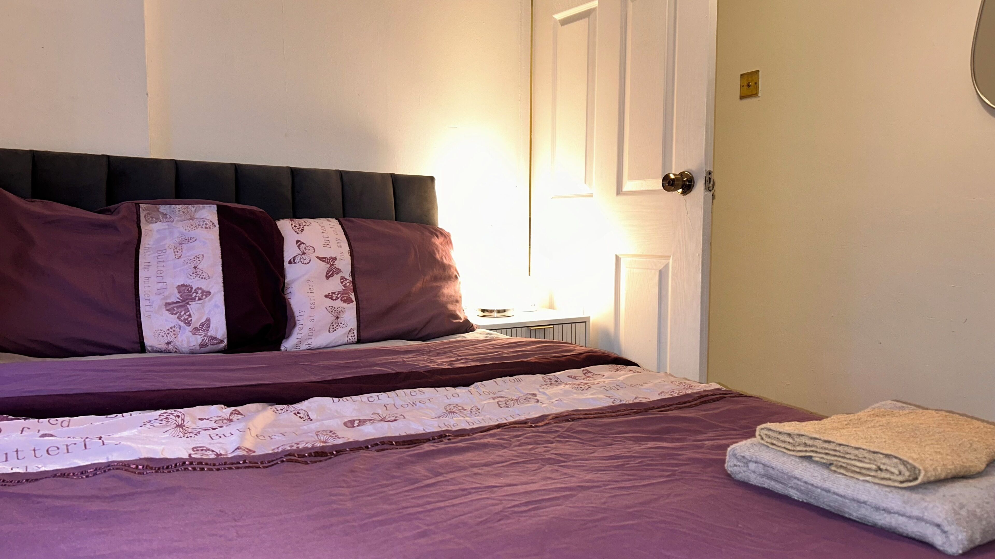 Un letto rifatto con una coperta viola e cuscini, un comodino con una lampada, e una porta con maniglia.