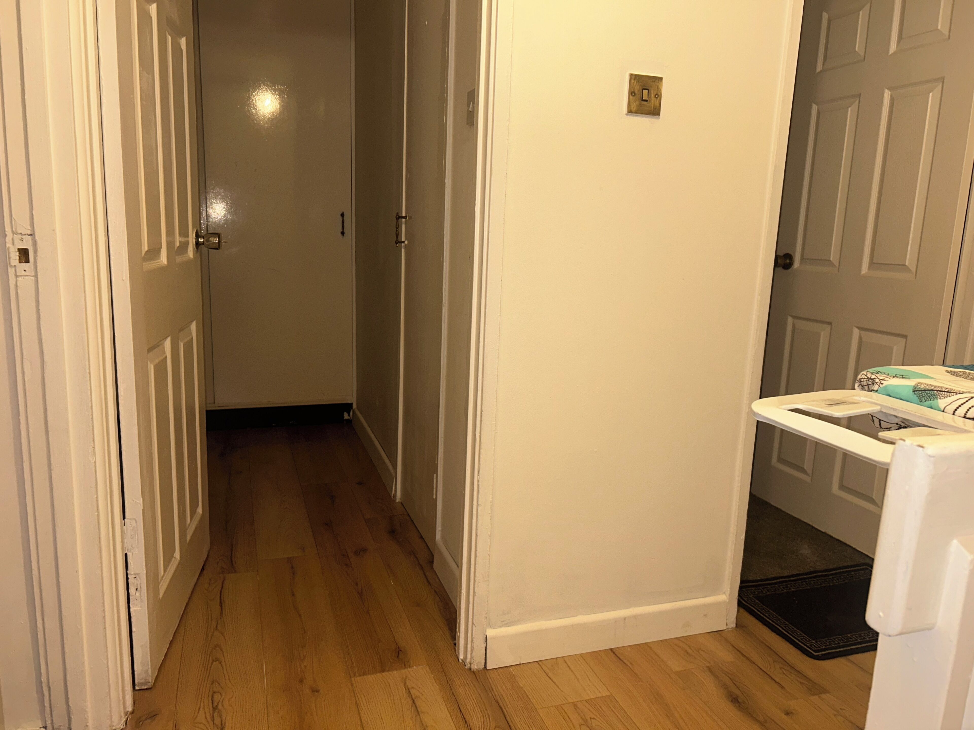A hallway with wooden flooring, white walls, and a door with a light switch.