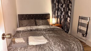 A neatly made bed with a patterned bedspread, a bedside lamp, and a radiator. - Charming 4-bedroom House Rental in Basildon, UK (Basildon)