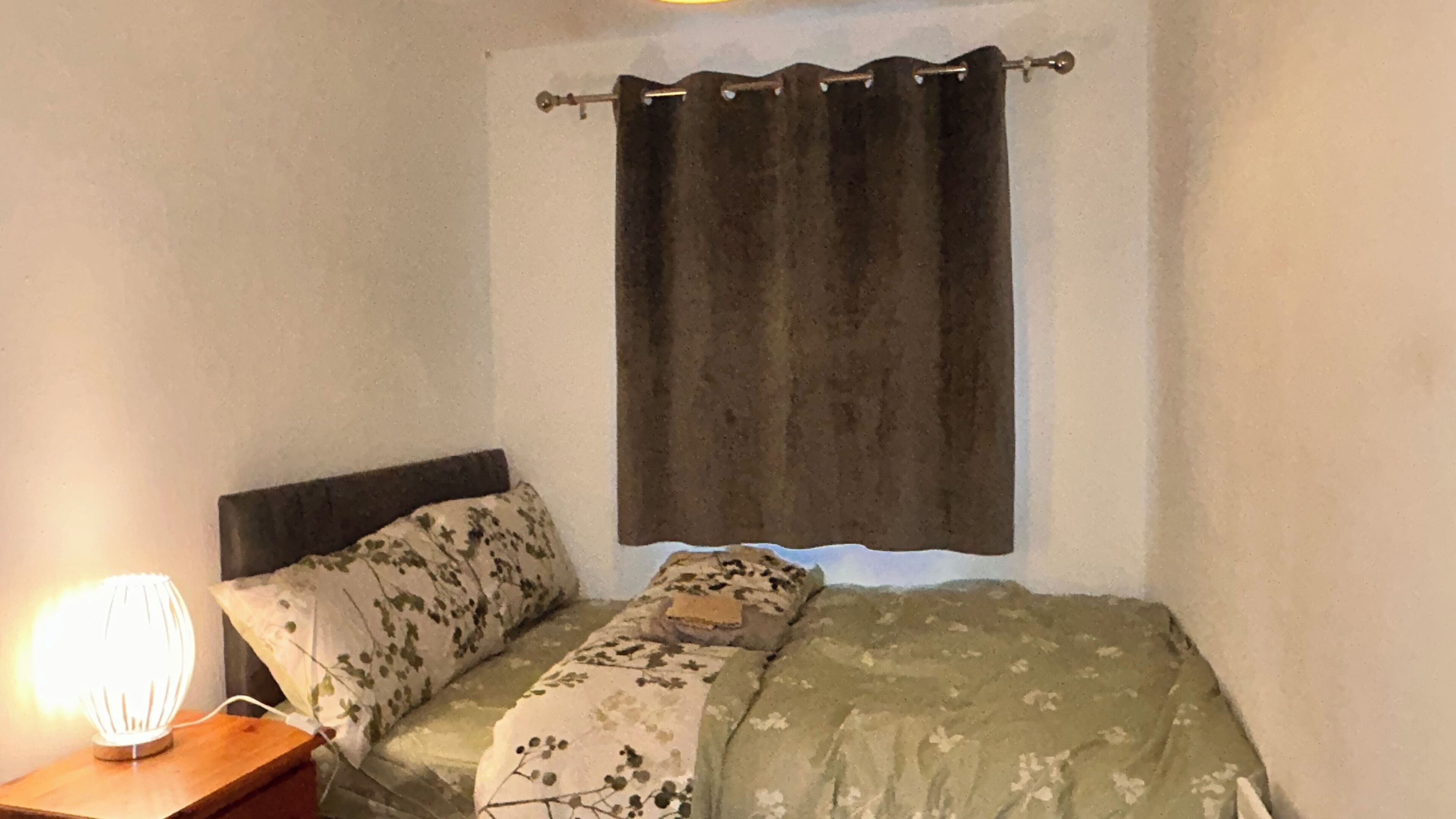 Ein kleines Zimmer mit einem Einzelbett, einem Nachttisch aus Holz, einer Lampe und einer Bettdecke mit Blumenmuster.