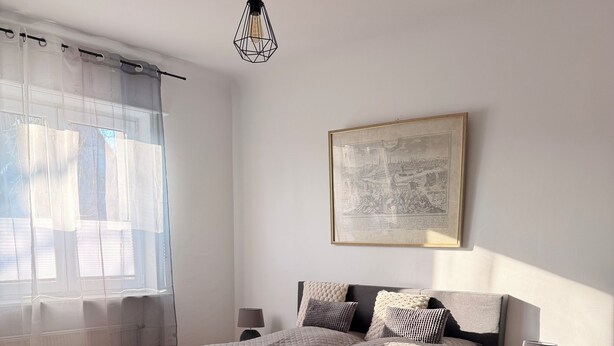 Ein Schlafzimmer mit einem großen Bett, einem Nachttisch mit Lampe, einem gerahmten Bild an der Wand und einem Fenster mit durchsichtigen Vorhängen.