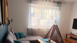 Ein Wohnzimmer mit grauem Sofa, einem Holztisch, einer Gitarre, einer zeltartigen Konstruktion und einem Fernseher auf einem Holzgestell.