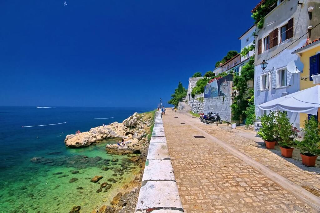 A coastal street with stone pavement, buildings with balconies, and a clear blue sea.