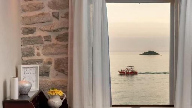 A room with a stone wall, a wooden desk with a vase and picture frame, a white chair, and a window with a view of the sea and a boat. - Dimore Apartment Monte (Rovinj)