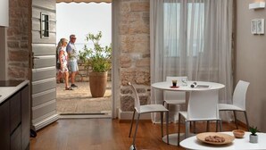A modern kitchen with a stone wall, a dining area with a round table and chairs, and a view of the sea through an open door. - Dimore Apartment Monte (Rovinj)