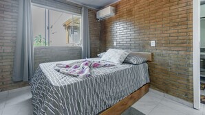 A bedroom with a brick wall, a bed with striped bedding, a window with curtains, and an air conditioning unit. - Apto Perto da Praia Com Piscina e Churrasqueira (Penha)