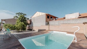A swimming pool with a wooden deck, a lounge chair, and a brick wall. - Apto Perto da Praia Com Piscina e Churrasqueira (Penha)