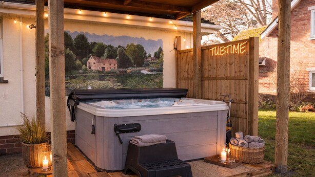 A hot tub in a wooden outdoor setting with a 'TUBTIME' sign, a wooden deck, and a backdrop of a painted landscape.