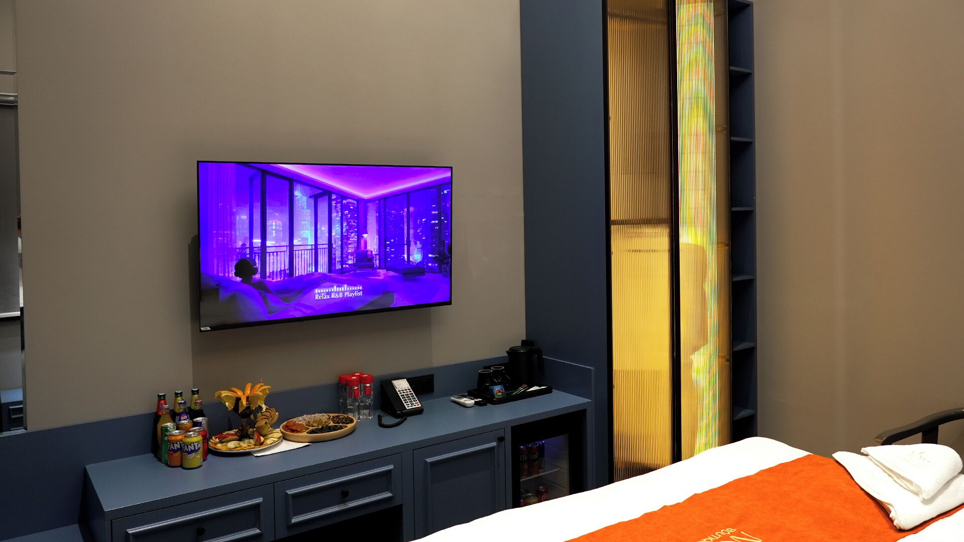 A hotel room with a flat-screen TV displaying a cityscape, a minibar with beverages, a tray of snacks, and a bed with an orange blanket.
