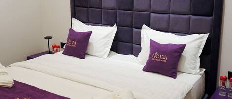 A hotel room with a large bed, two pillows with the hotel's logo, a purple throw blanket, and a nightstand with a lamp and bottles.