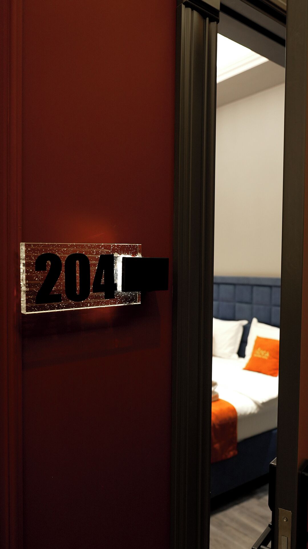 Een hotelkamerdeur met een nummerbord '204' en een blik op een bed met kussens en een oranje accent.