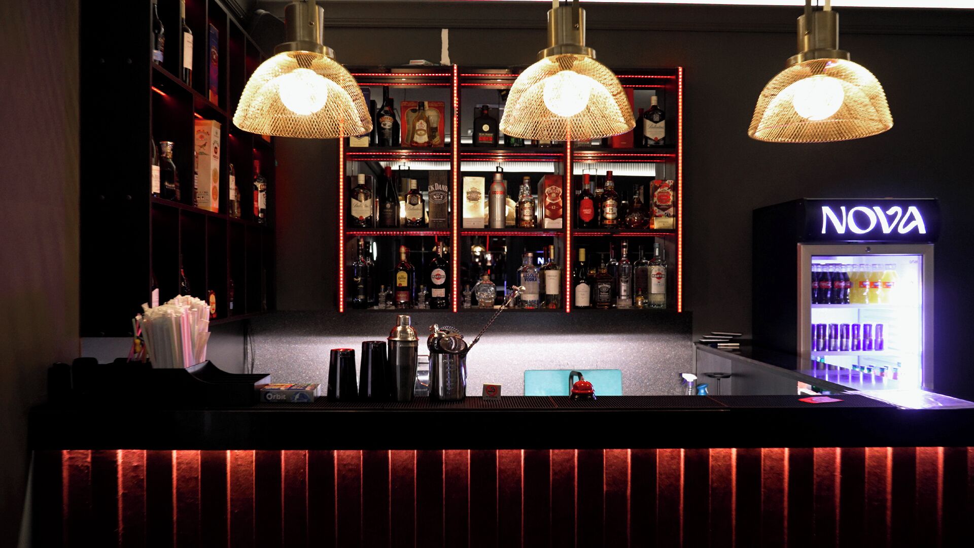 A bar area with shelves of liquor bottles, a NOVA branded refrigerator, and three hanging lamps.