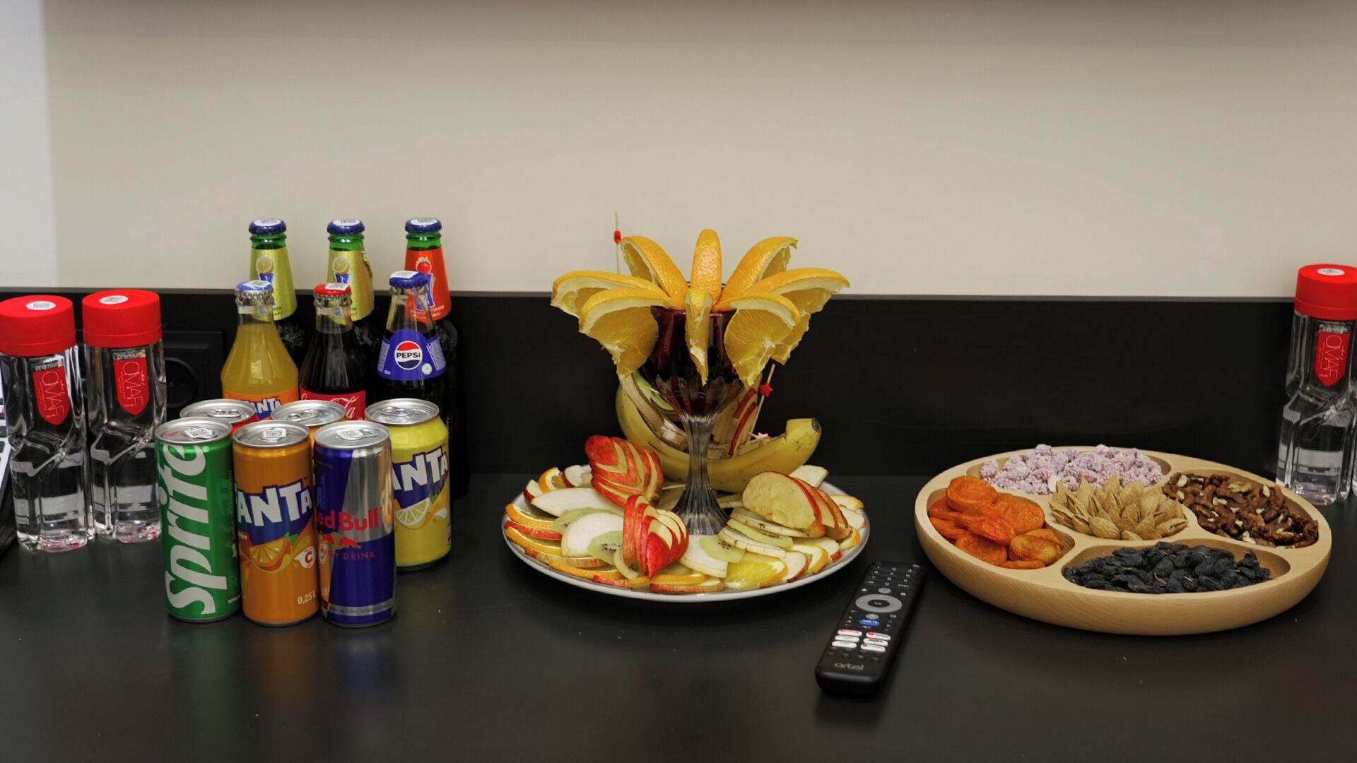 A table with beverages, snacks, a remote control, and a decorative fruit arrangement.