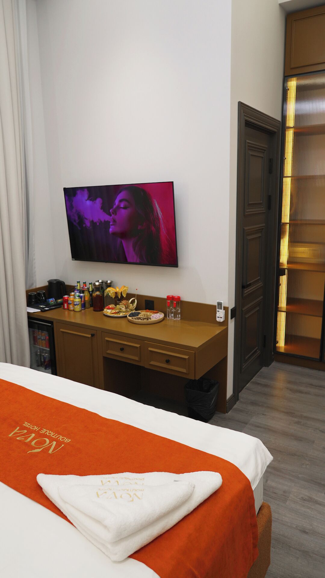 A hotel room with a bed, a desk with a TV, a minibar, and a door with a lighted panel.