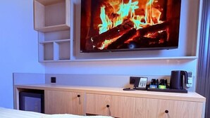 Habitación de hotel con cama, televisor de pantalla plana mostrando una chimenea, minibar y escritorio con cafetera.