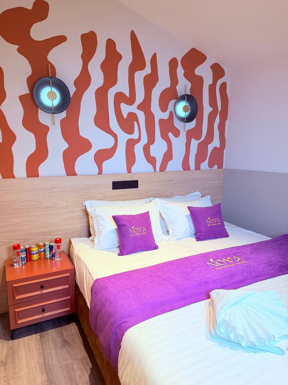 A hotel room with a bed, two wall-mounted speakers, a nightstand with a lamp, and a minibar.