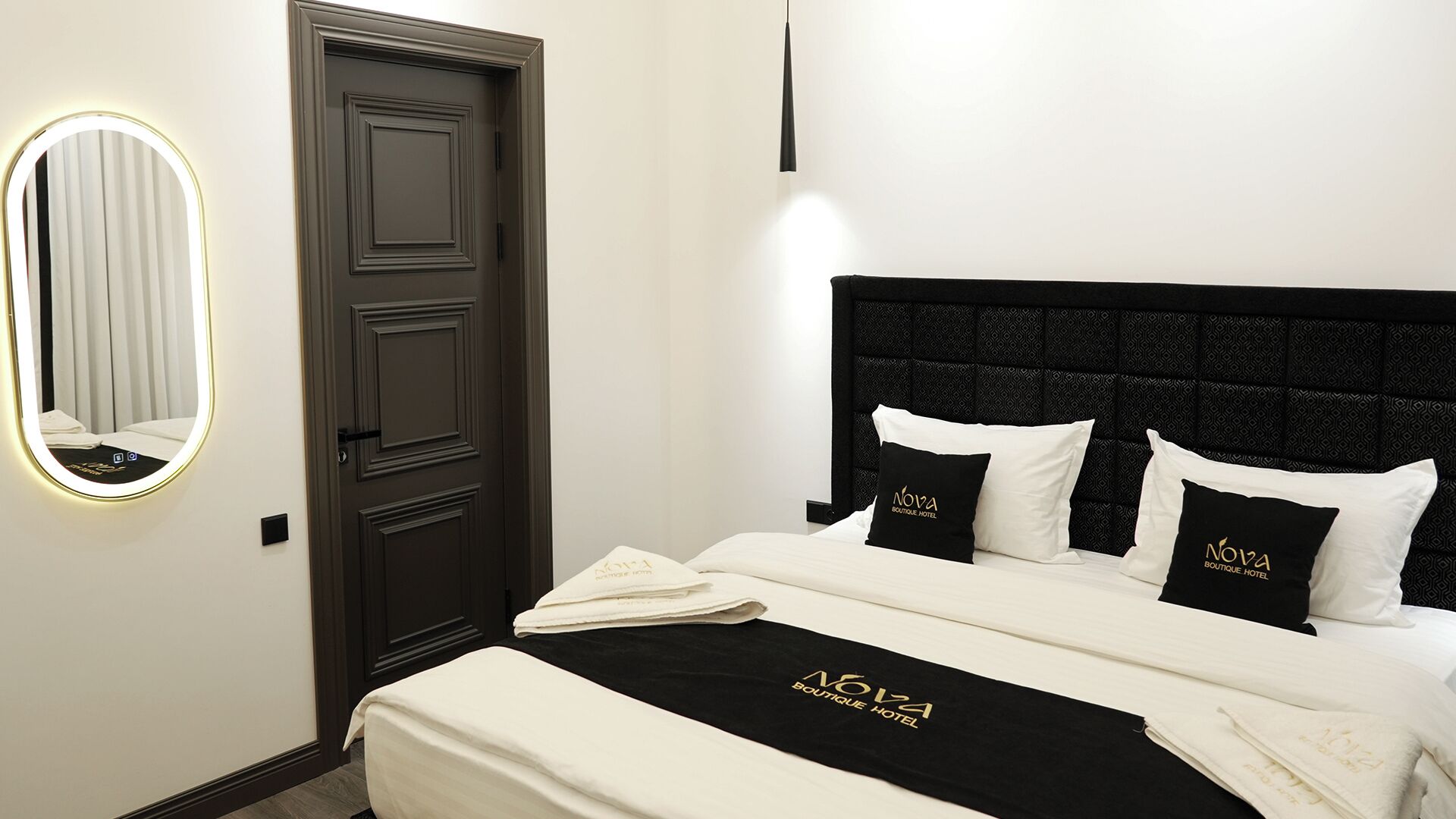 Habitació d'hotel amb una porta de fusta fosca, un mirall rodó amb il·luminació, un llit amb roba blanca i almoines negres i blanques, i un panell frontal de llit negre.