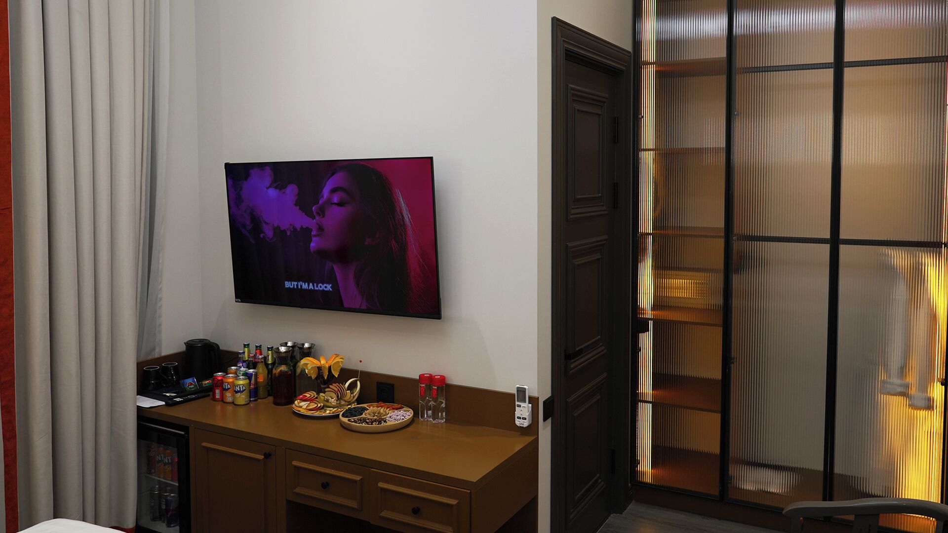 A hotel room with a flat-screen TV, a minibar, a desk with snacks, and a sliding door.